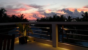 A balcony with a view of a sunset over a body of water, palm trees, and a railing.