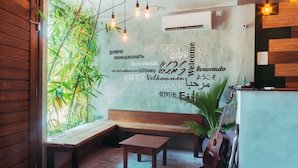 A reception area with a wooden bench, a small table, and a guitar. There is a wall with multilingual welcome signs and a potted plant.