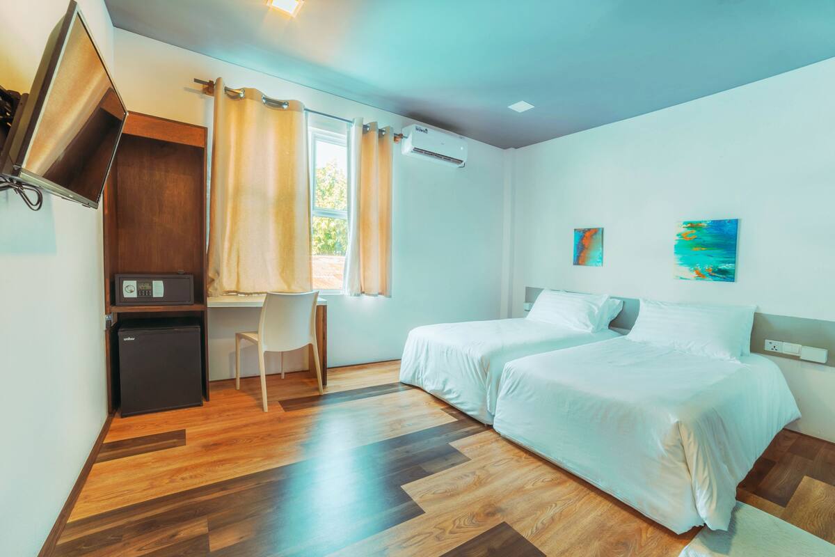 A hotel room with a desk, two beds, a TV, and an air conditioning unit.