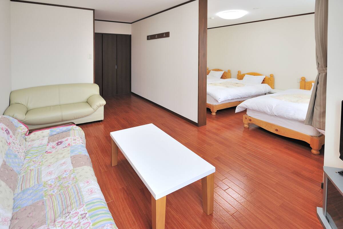 A hotel room with a sofa, two beds, a white table, and wooden flooring.
