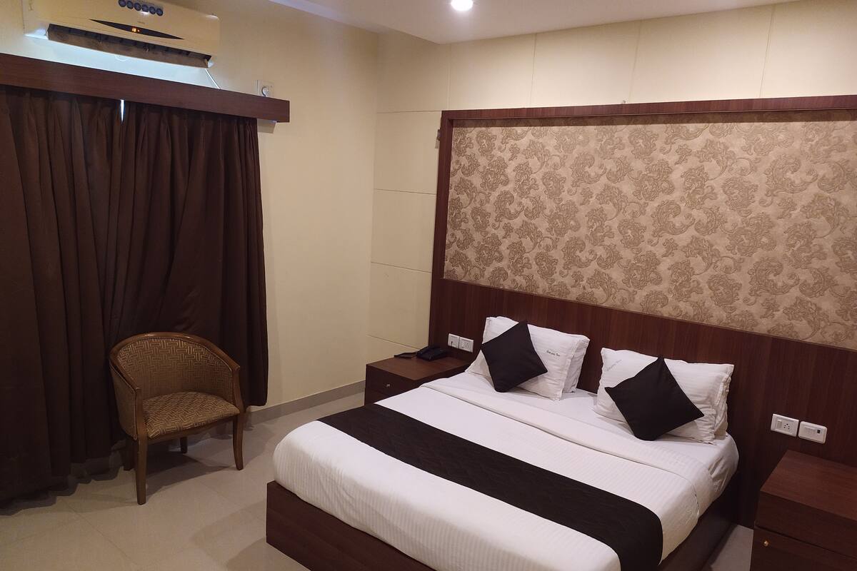 A hotel room with a bed, a chair, a nightstand, and a wall-mounted air conditioner.
