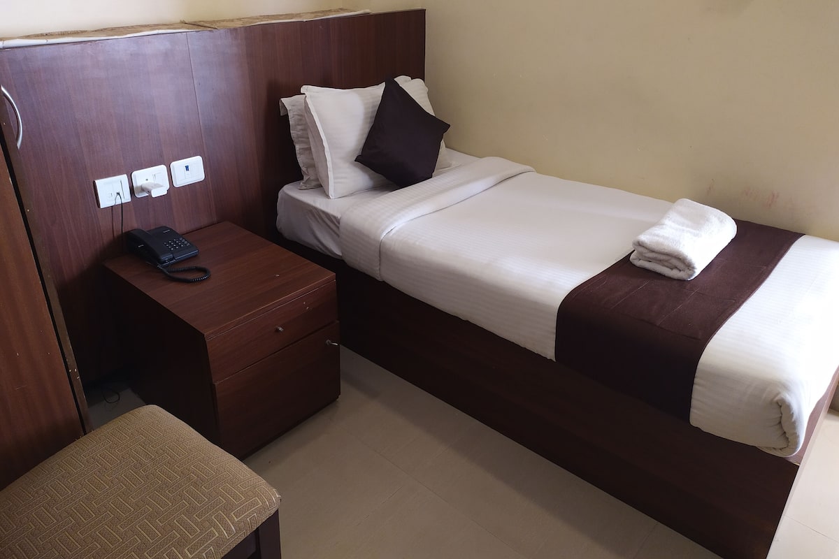 A single bed with a brown headboard, a bedside table with a phone, and a power outlet on the wall.