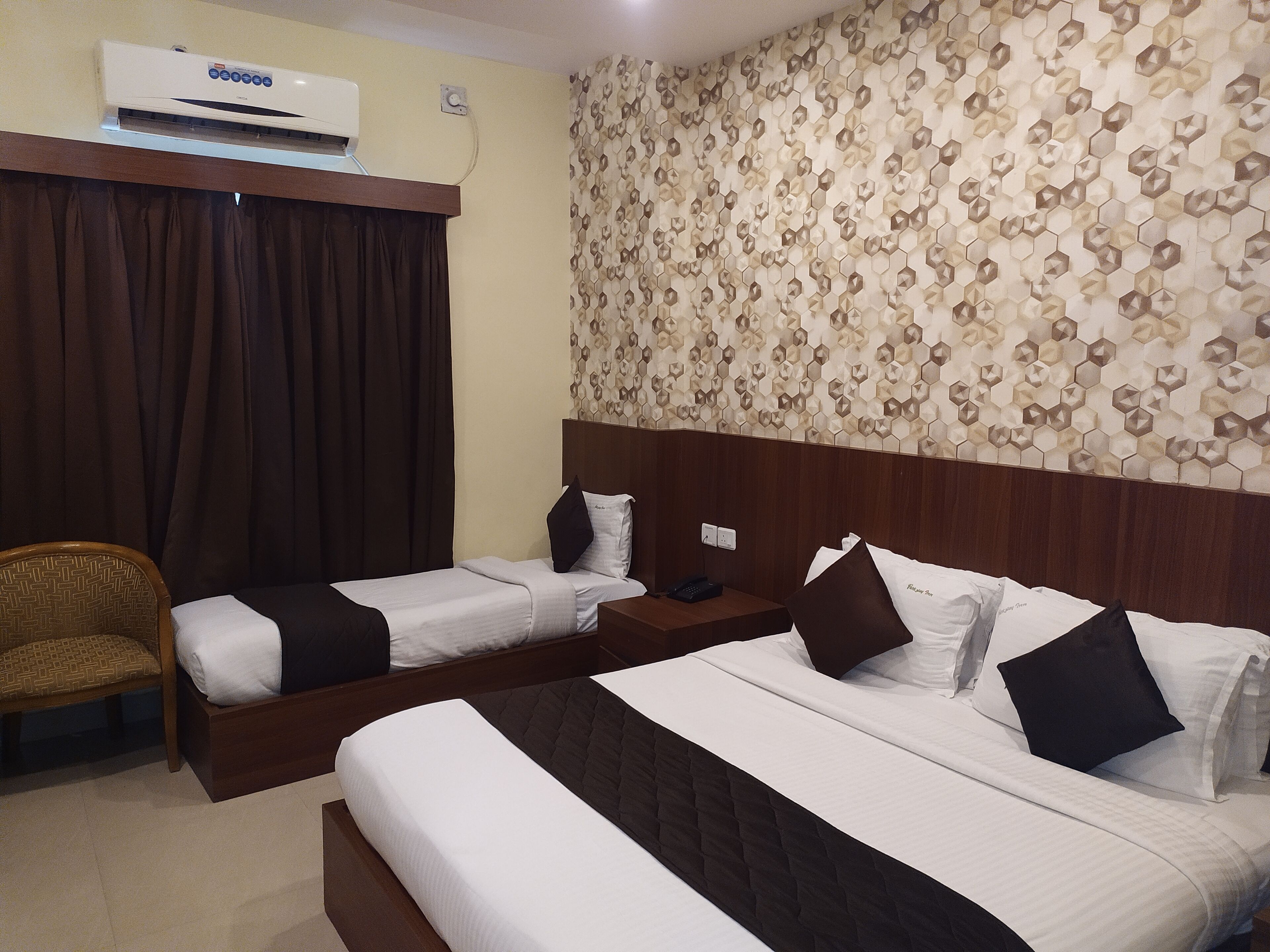A hotel room with two single beds, a wooden headboard, a wall air conditioner, a patterned wallpaper, and a brown curtain.