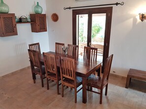 Uma área de refeições com uma mesa e cadeiras de madeira, uma pequena estante com objetos decorativos e uma porta que dá para uma área externa.