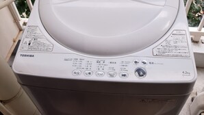 A white Toshiba washing machine with a control panel, located on a balcony with a metal railing and greenery in the background. - Elitz INN Third (Kyoto)