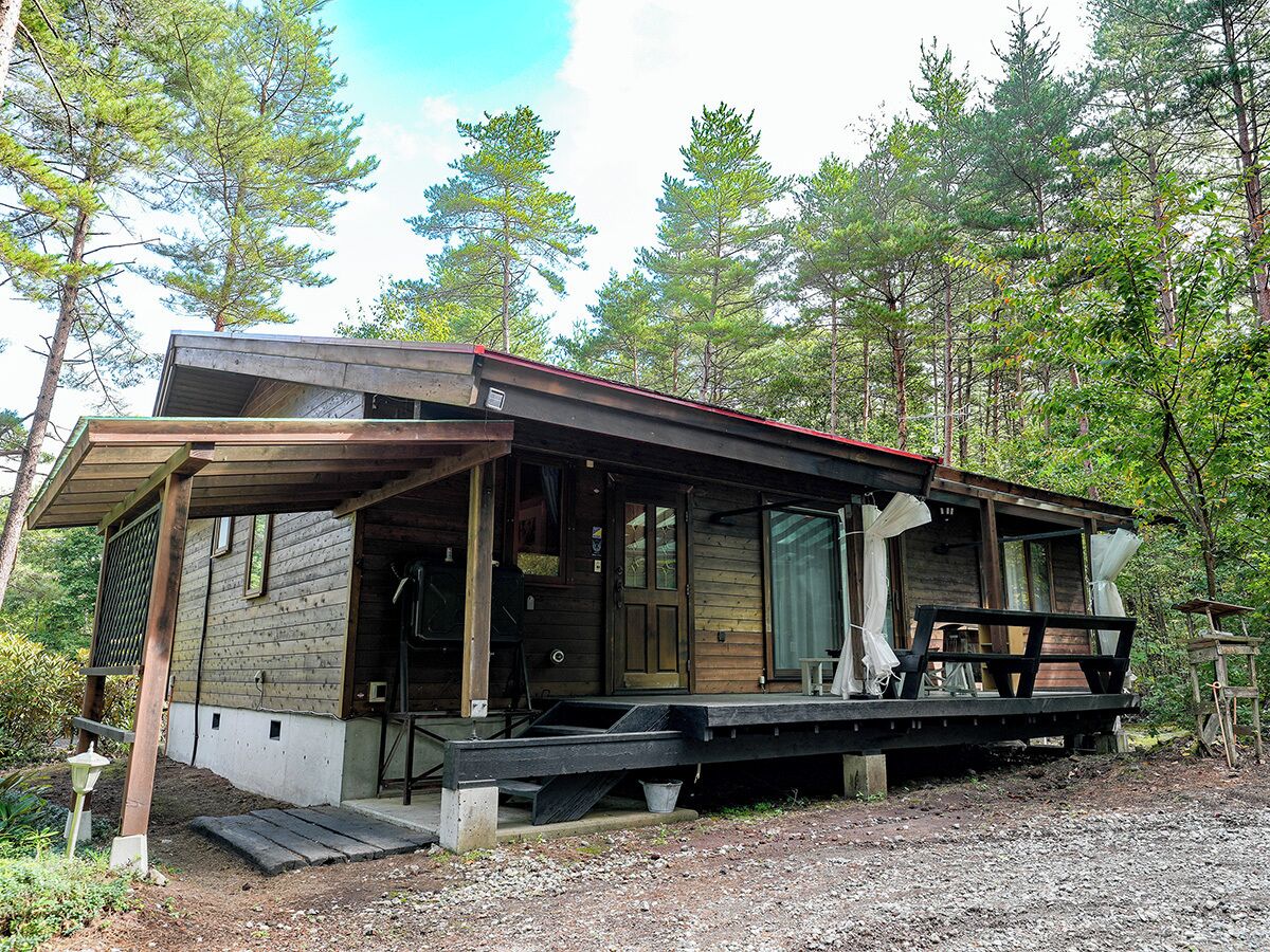 A rustic cabin with a porch, a covered deck, and a gravel driveway surrounded by trees.