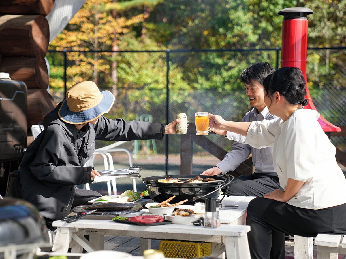 A group of people toasting with drinks at an outdoor dining area with a grill, surrounded by trees.
