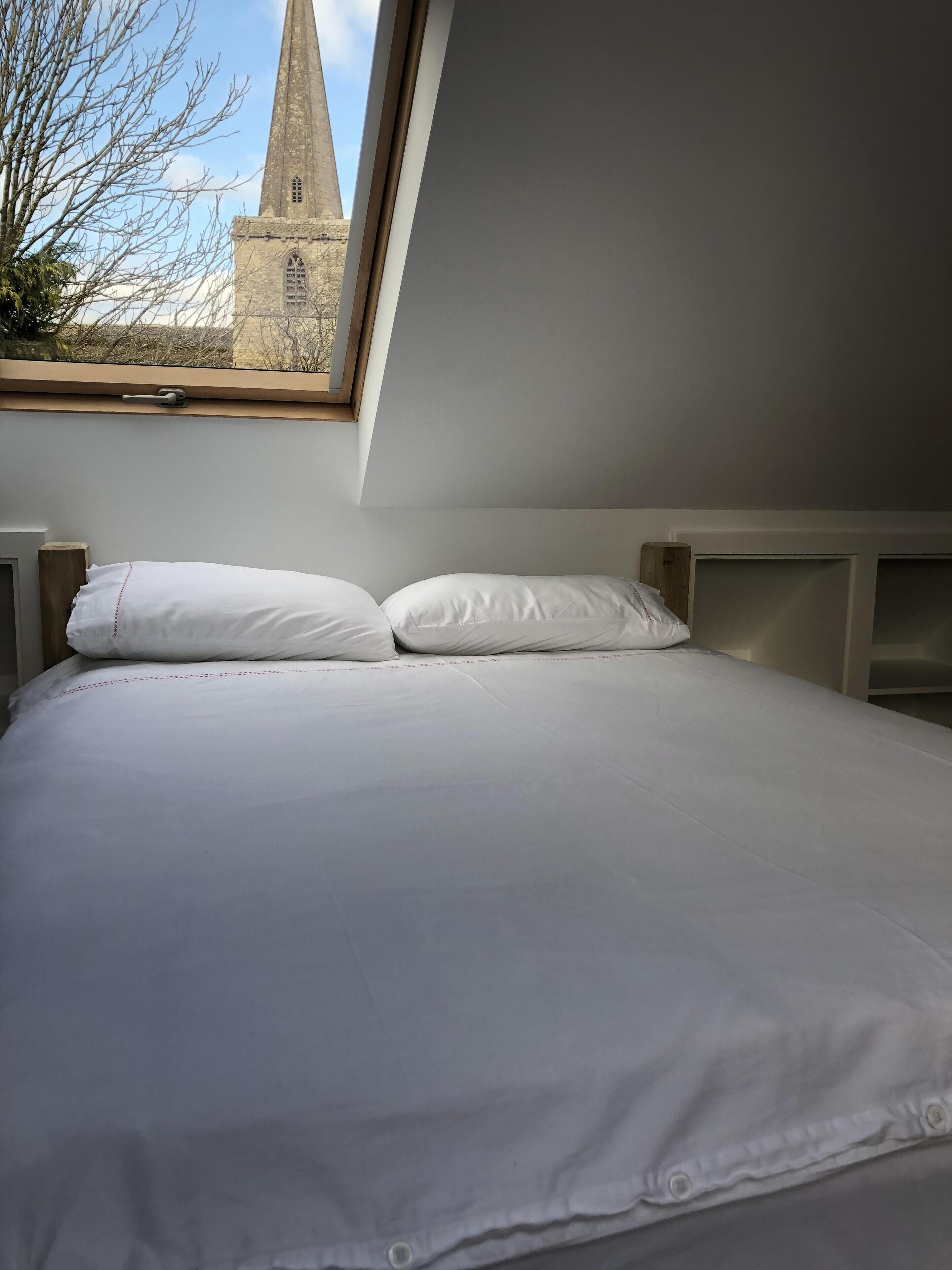 A bedroom with a bed, white bedding, a window with a view of a church spire, and a wooden headboard.