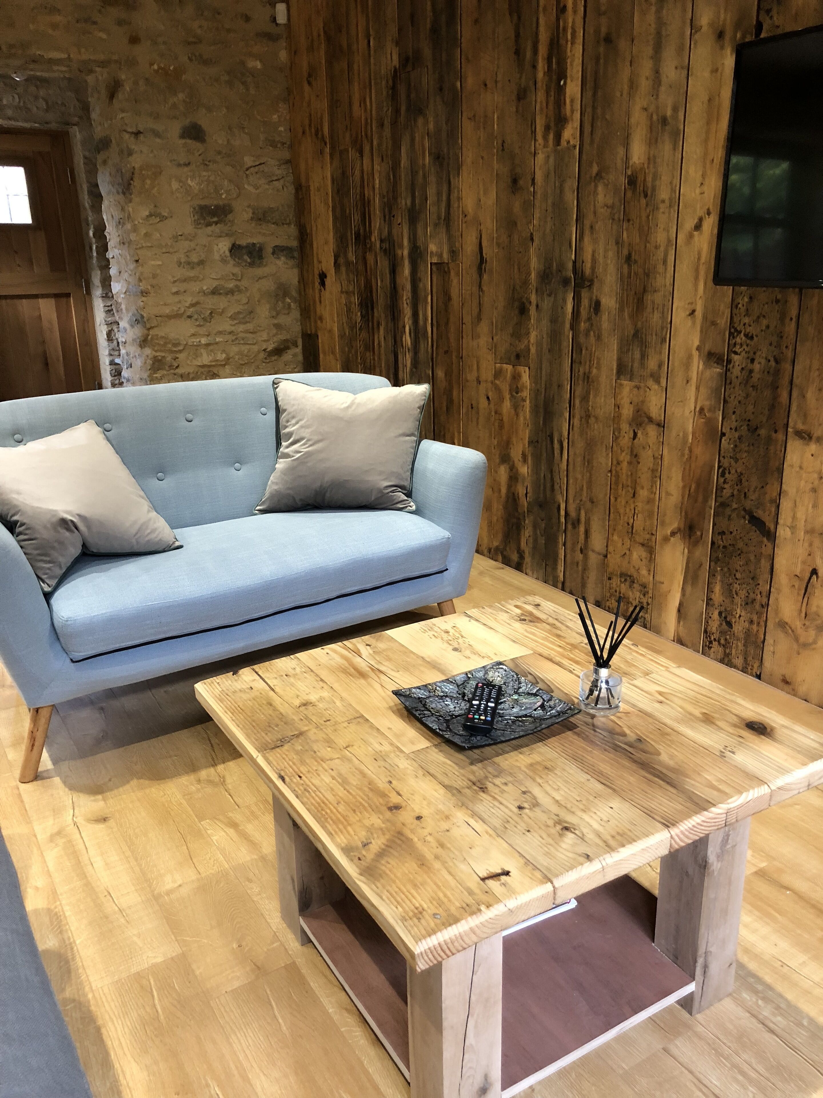 Una habitación con un sofá azul claro frente a una pared de madera, una mesa de centro de madera con un control remoto y un recipiente de vidrio, y un televisor montado en la pared.