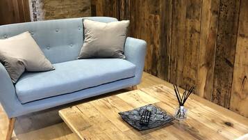 Una habitación con un sofá azul claro frente a una pared de madera, una mesa de centro de madera con un control remoto y un recipiente de vidrio, y un televisor montado en la pared.