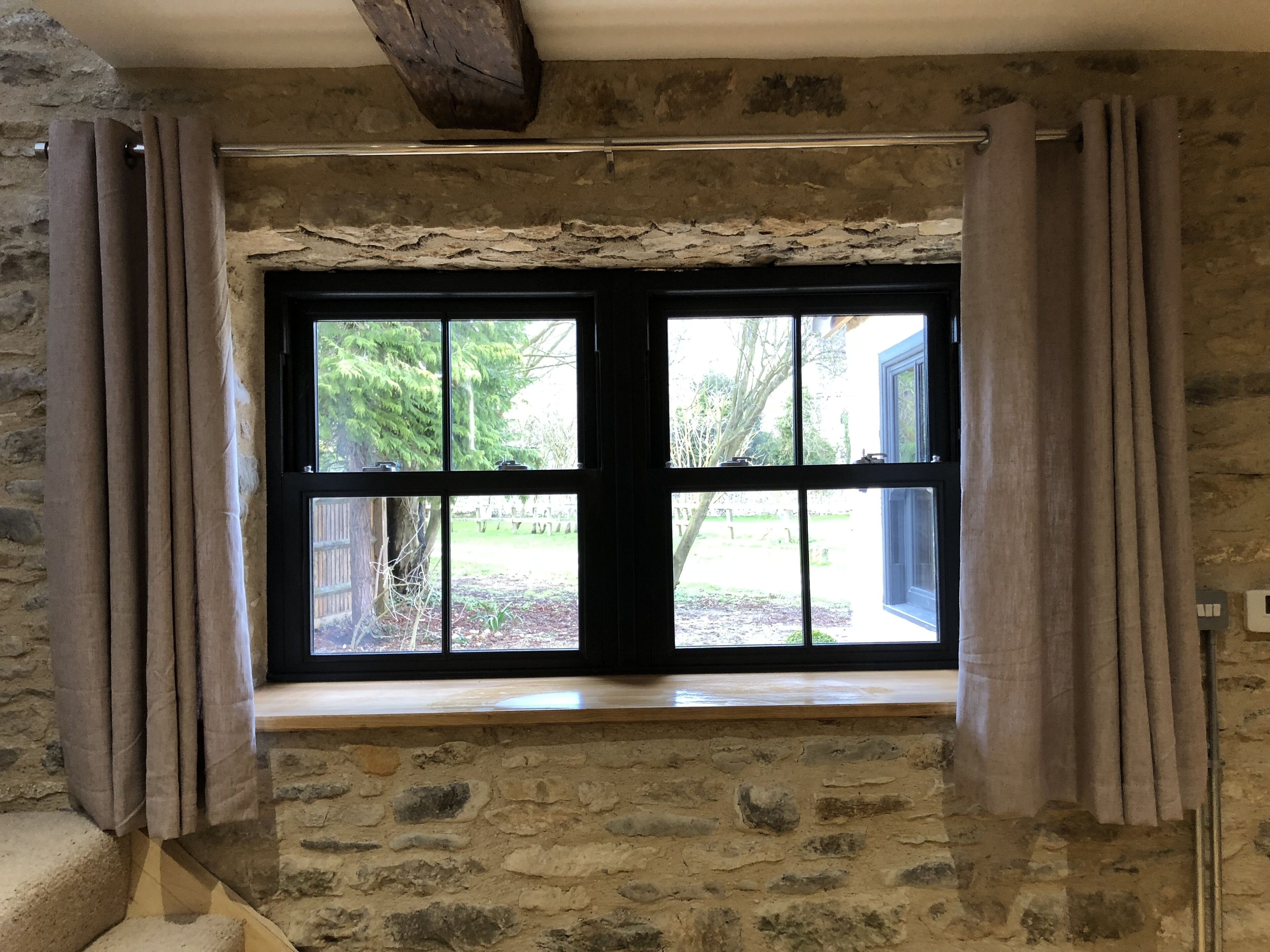 Ein Raum mit Steinwänden, einem großen Fenster, Vorhängen und einer Holzbalkendecke.