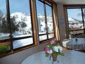 A room with large windows offering a view of snow-covered mountains and a ski slope. There is a table with a vase of flowers and a calendar on the wall. - Ouji Penshon Naeba (Yuzawa)