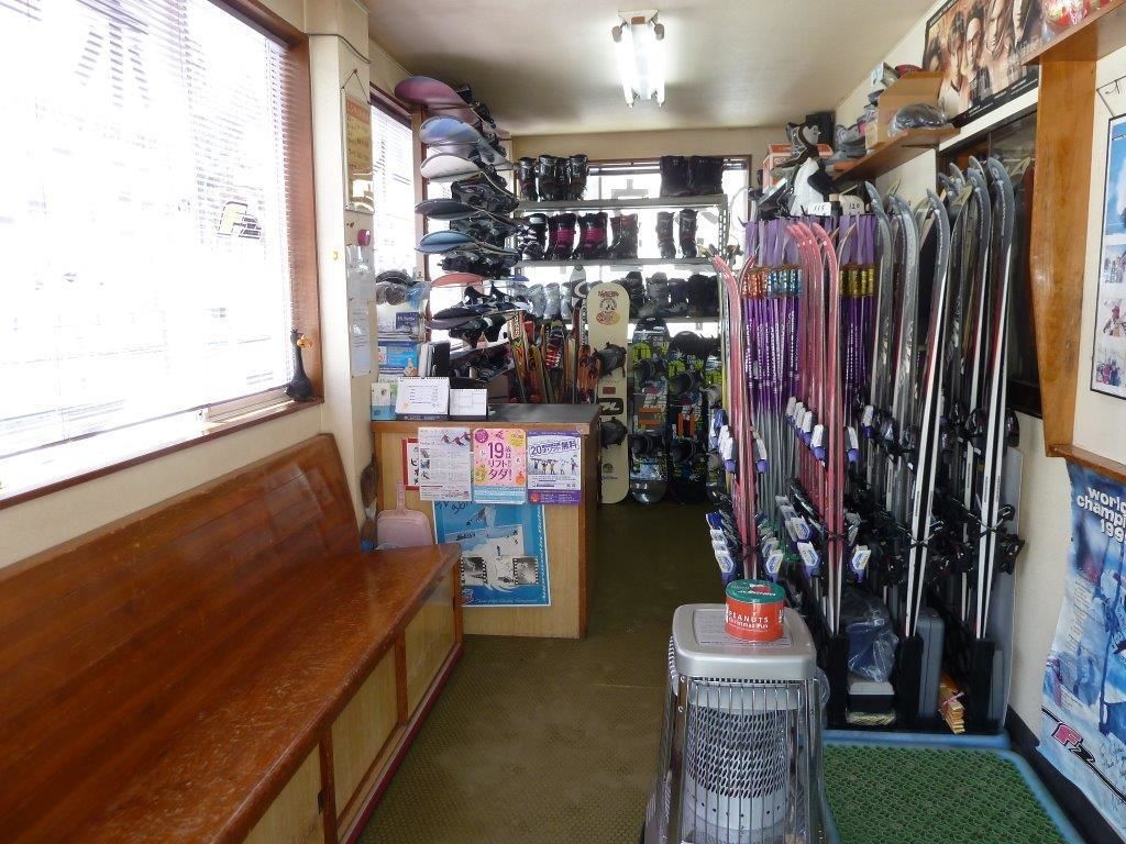 A narrow shop with shelves of skis and snowboards, a wooden bench, and a small stool with a container.