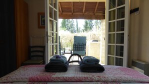 Une chambre avec une porte ouverte donnant sur un patio. On y voit un lit recouvert d’une courtepointe à motifs floraux et deux couvertures pliées. Une chaise et une table sont visibles à l’extérieur de la porte.