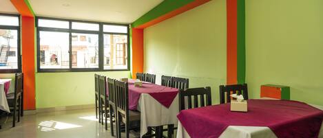 A dining area with tables covered in white and maroon tablecloths, wooden chairs, and a window with a view of buildings outside.