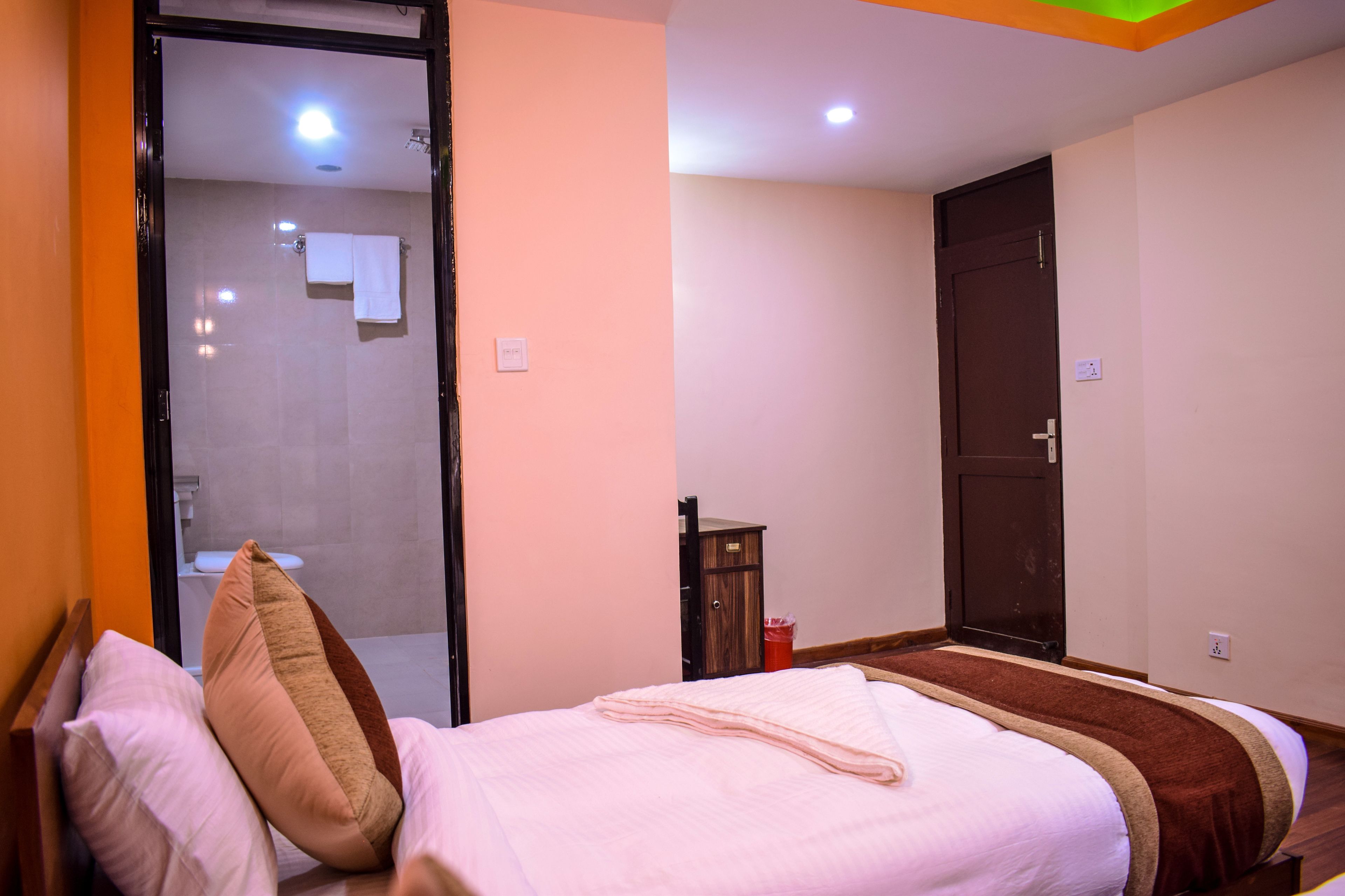 A hotel room with a bed, a wooden wardrobe, a bathroom visible through an open door, and a small table with a red trash bin.