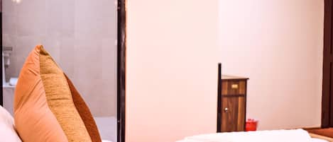 A hotel room with a bed, a wooden nightstand, and a bathroom visible through a glass door.