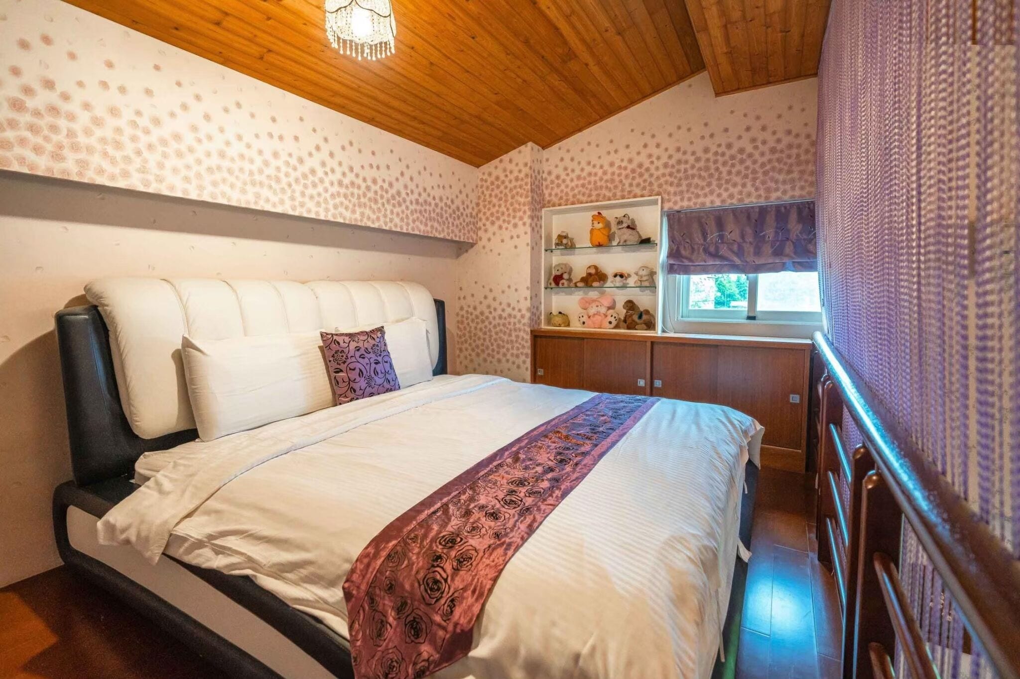 Un dormitorio con una cama, cabecera, estante con peluches y una ventana con cortinas.