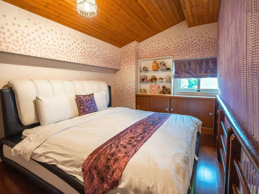 Un dormitorio con una cama, cabecera, estante con peluches y una ventana con cortinas.