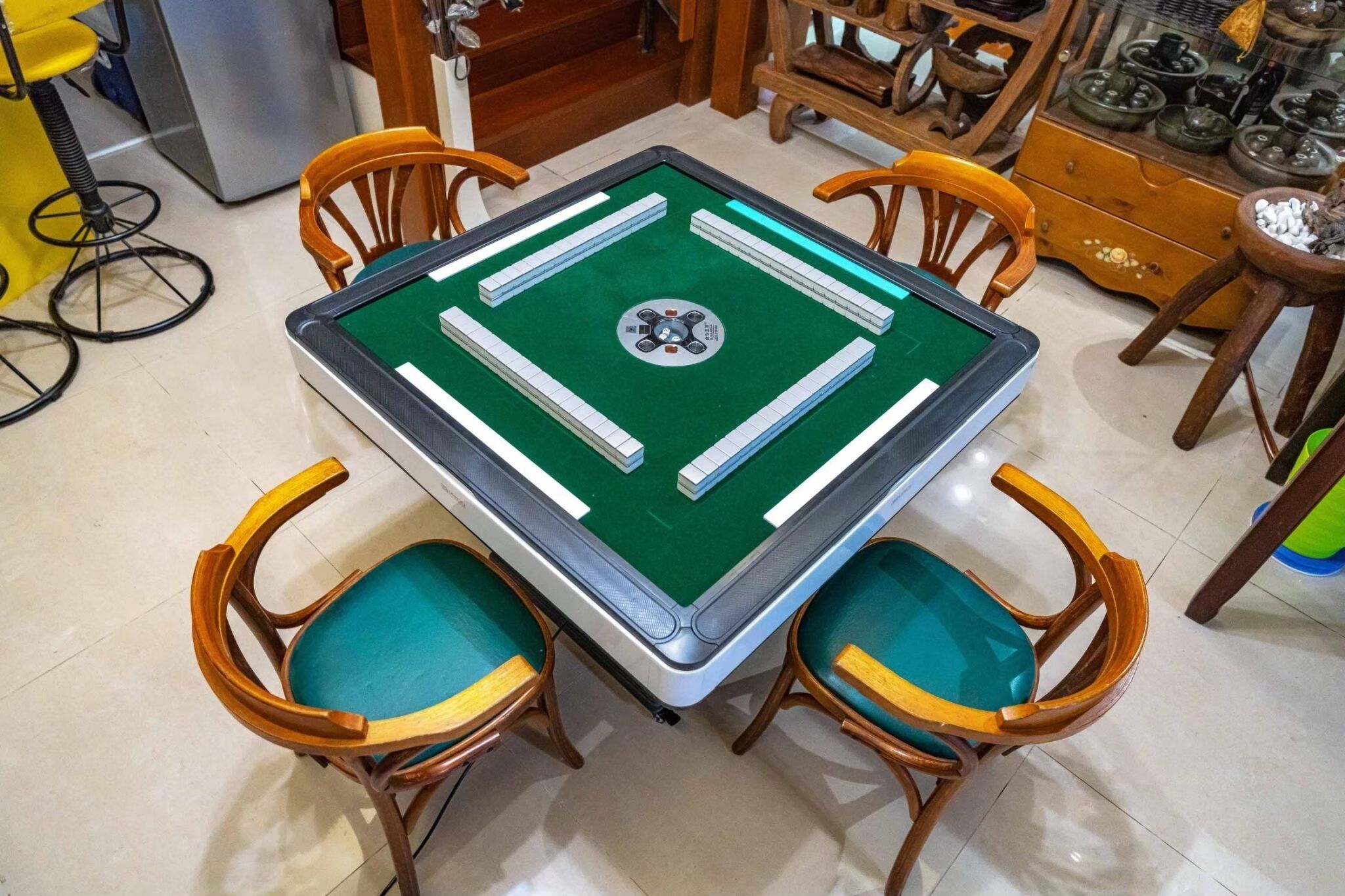 A square pool table with a green felt surface, surrounded by four wooden chairs with teal cushions.