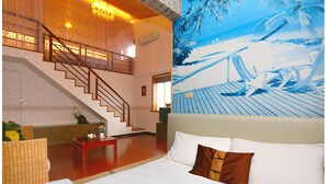 Quarto de hotel com uma cama, uma mesa vermelha, uma escada e um grande mural de praia na parede.
