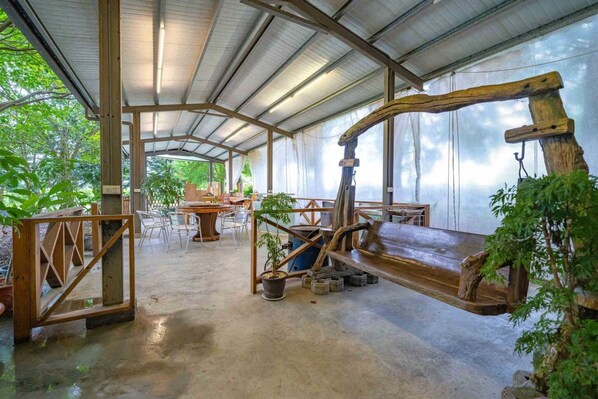 Un espacio al aire libre techado con un banco de madera rústica, mesas, sillas y plantas en macetas.