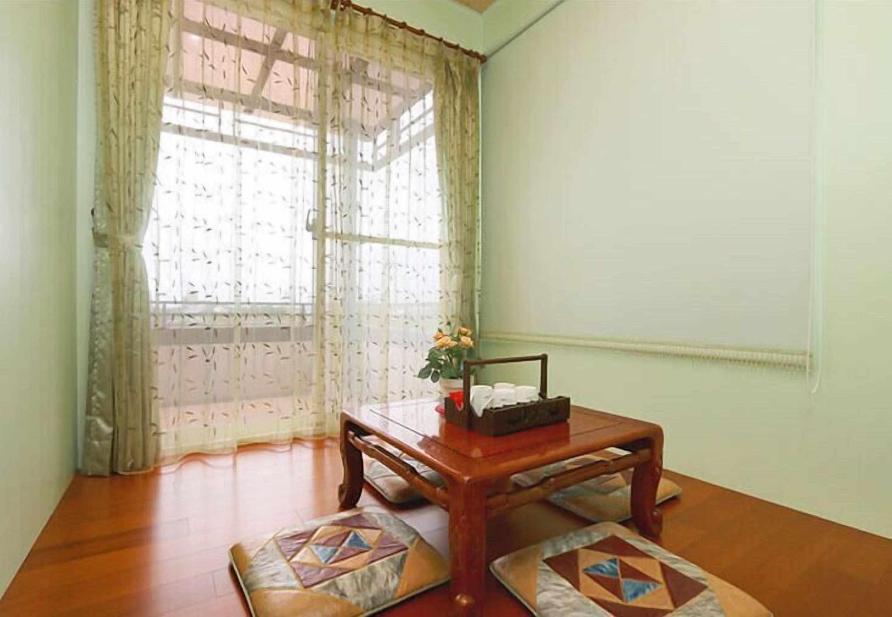 A room with a wooden table, two patterned cushions, a small plant, and a window with sheer curtains.