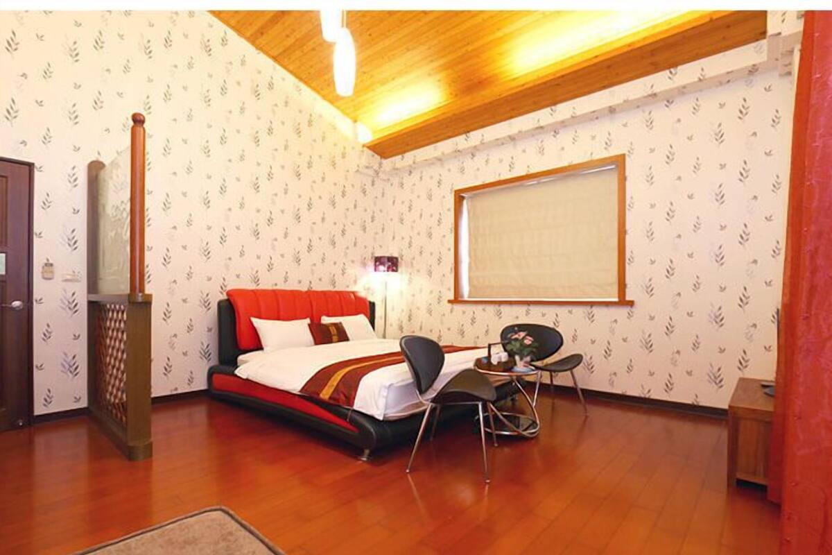 A hotel room with a bed, a desk with two chairs, a window, and a wooden ceiling.