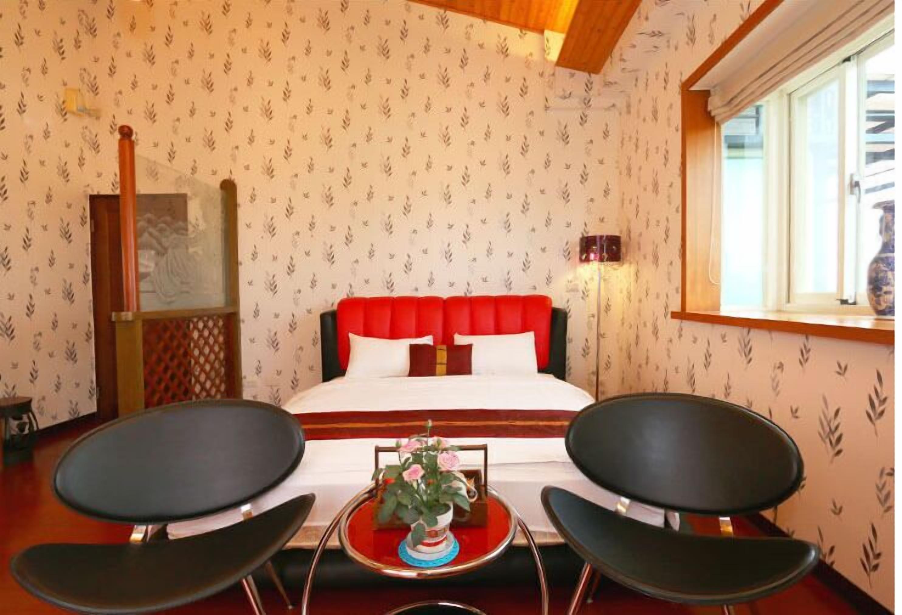 A hotel room with a bed, two black chairs, a small table with a vase of flowers, and patterned wallpaper.