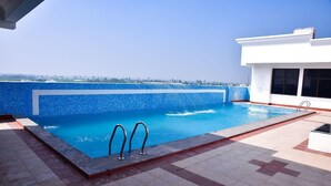 A rooftop swimming pool with clear blue water, surrounded by a tiled deck and a white building.