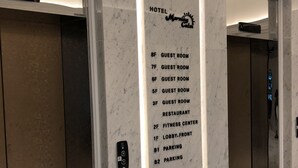Un vestíbulo de hotel con piso y paredes de mármol, que incluye un ascensor con pantalla digital y un directorio en la pared que enumera habitaciones de huéspedes, restaurante, gimnasio, área de recepción y estacionamiento.