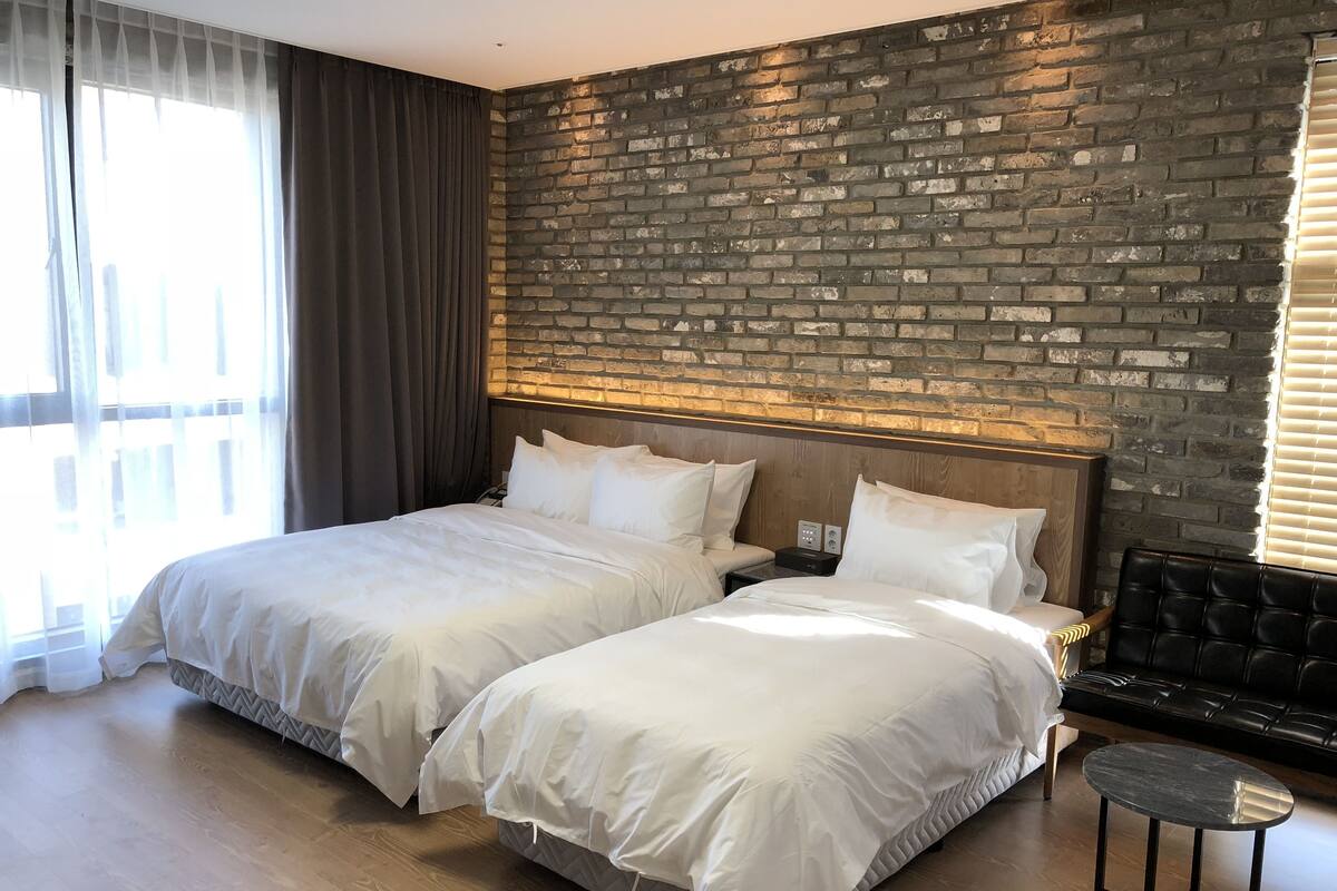 A hotel room with two beds, a brick accent wall, a wooden floor, and a black leather sofa.