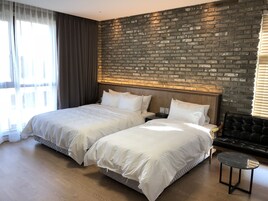 Quarto de hotel com duas camas, parede de tijolos decorativa, piso de madeira e sofá de couro preto.