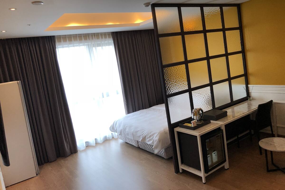 A hotel room with a bed, a desk, a chair, a small table, and a refrigerator.