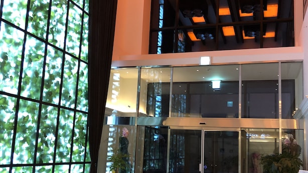Un vestíbulo moderno con un amplio acceso por puerta de vidrio, una zona de estar con sofás y una mesa de centro, y un techo con vigas a la vista e iluminación empotrada.