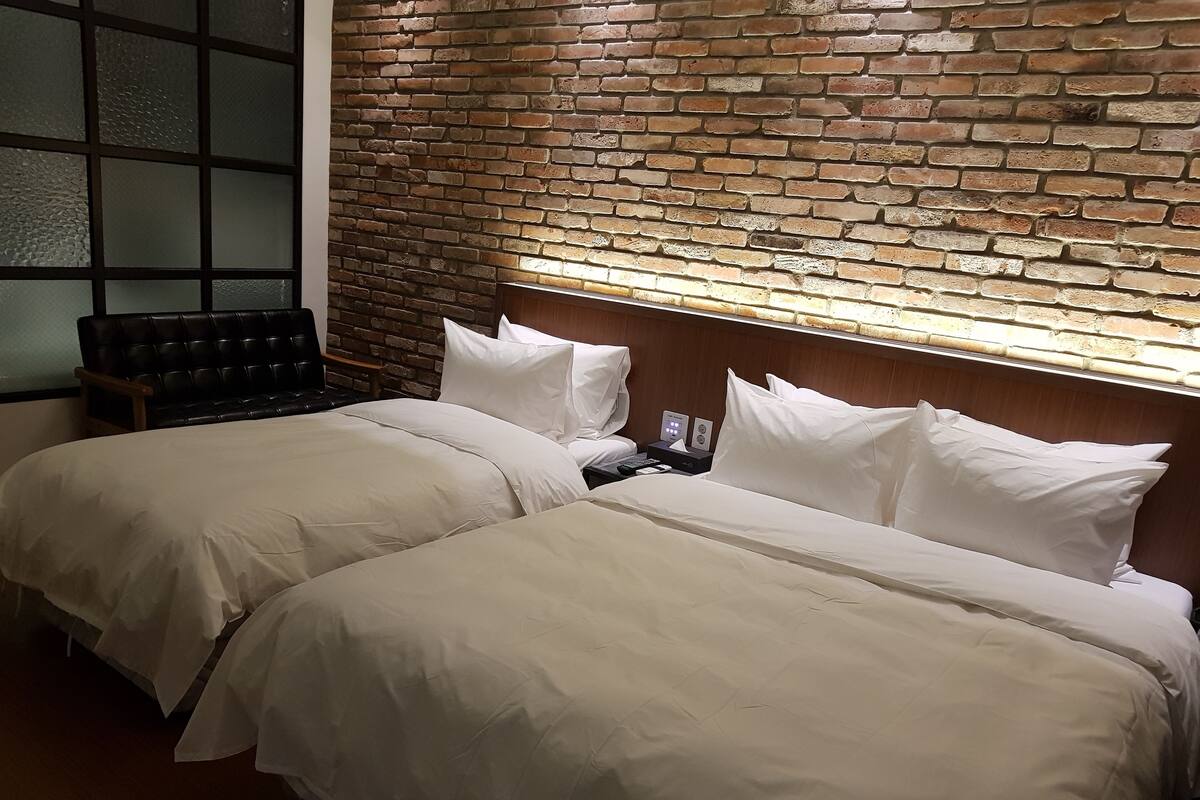 A hotel room with two beds, a brick wall, a wooden headboard, and a window with frosted glass.