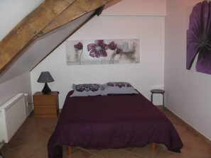 A bedroom with a sloped ceiling, a bed with a purple bedspread, two pillows, a nightstand with a lamp, and a wall-mounted flower painting. - house / villa - PONTIGNE (PONTIGNE)