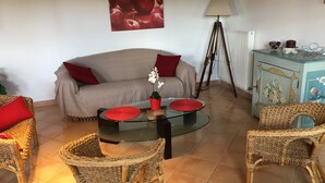 Una sala de estar con un sofá, dos sillones de mimbre, una mesa de centro de vidrio, una lámpara de pie y un mueble con motivos florales.