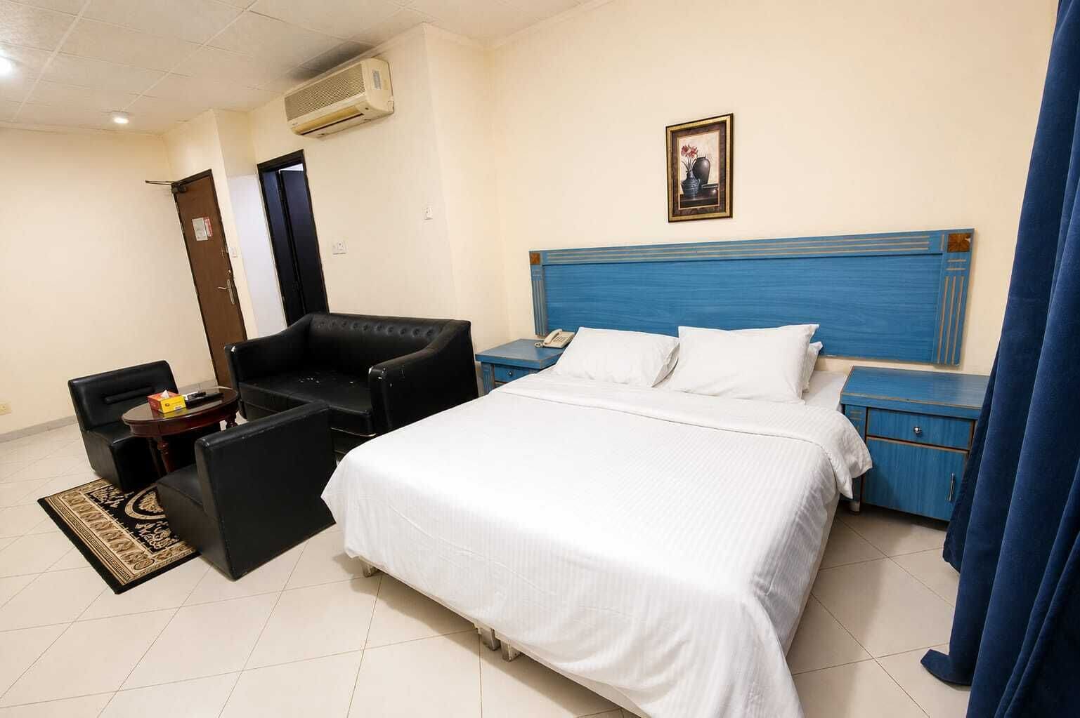 A hotel room with a bed, a desk, a chair, a nightstand, a picture on the wall, and an air conditioning unit.