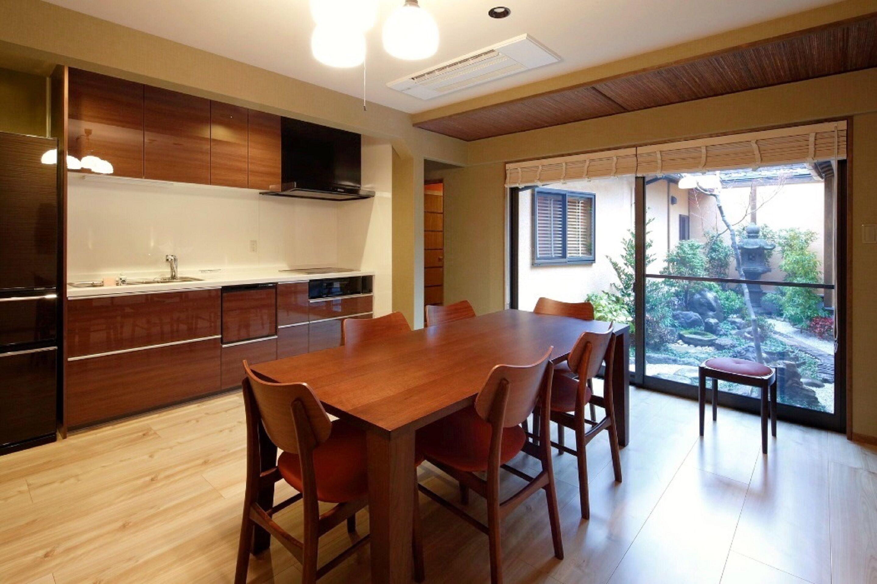 A modern kitchen with wooden cabinets, a large dining table, and a view of a garden through large windows.