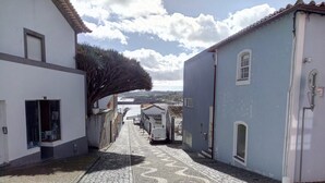 A cobblestone street with buildings on either side, a clear sky, and a distant view of the sea. - Janelas da Praia, Apartment by the beach. (Praia da Vitória)