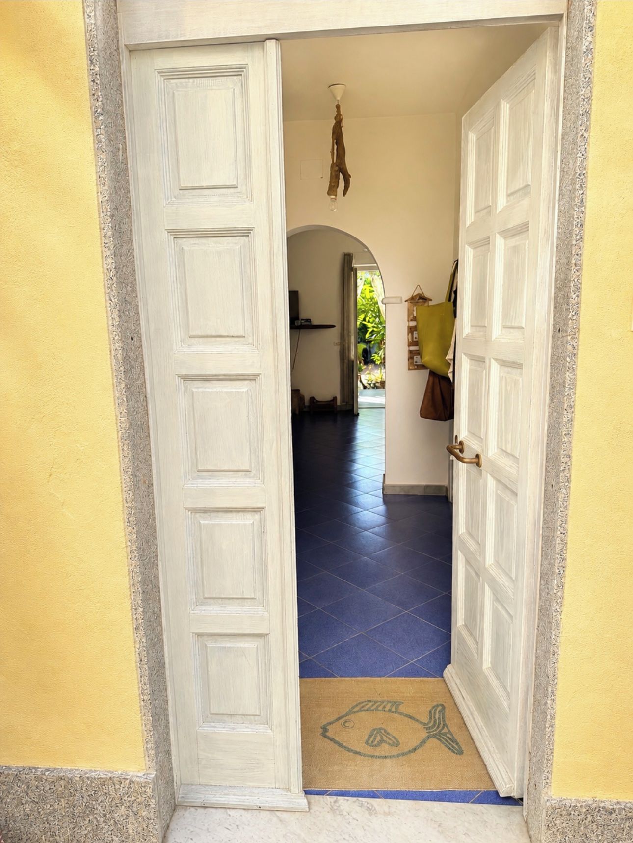 Un couloir avec une porte blanche sur la gauche et une porte blanche sur la droite, qui donnent sur une pièce dont le sol est recouvert de carreaux bleus.