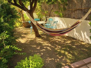 Un'amaca tra gli alberi in un giardino con tavolo e sedie, immersa nel verde e circondata da una staccionata di bambù.