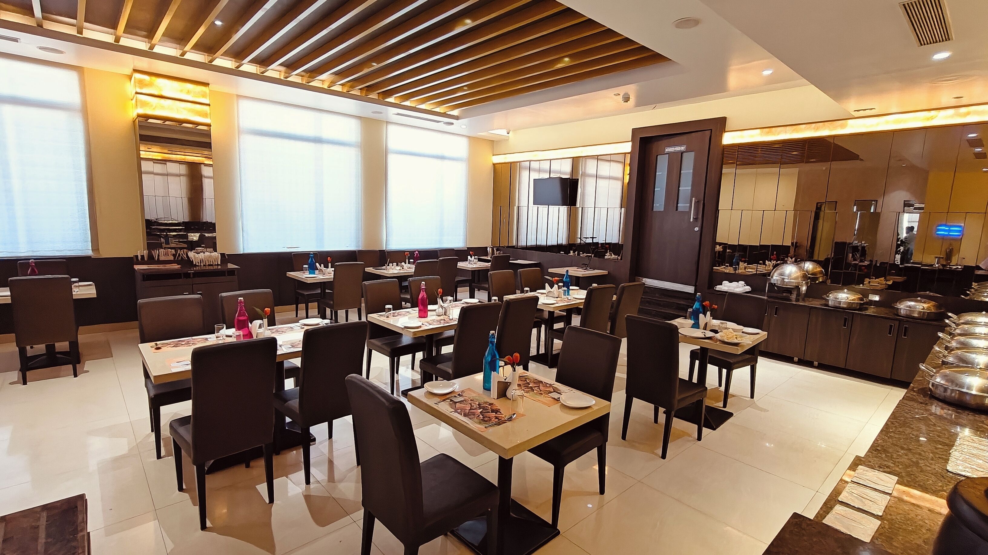 A modern restaurant with multiple dining tables and chairs, large windows, and a kitchen area visible in the background.