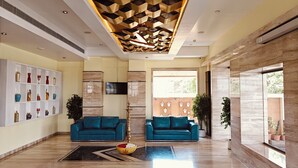 A modern lobby with two blue sofas, a decorative ceiling, a wall-mounted TV, and a large window. - ASR Parklane, A Bergamont Hotel - Hosur (Hosur)
