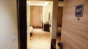 A hotel room with a wooden door marked with the number 407, a bed, a desk with a chair, and a mirror on the wall. - ASR Parklane, A Bergamont Hotel - Hosur (Hosur)