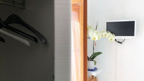 Ein Zimmer mit weißer Wand, einem an der Wand befestigten Fernseher, einem Holzrahmen für eine Tür, einem weißen Regal mit einer Pflanze und einem Kleiderständer mit schwarzen Kleiderbügeln.