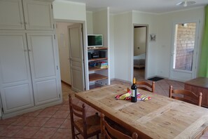 A dining area with a wooden table, chairs, and a bottle of wine. There is a TV on a shelf and a bed in the background.