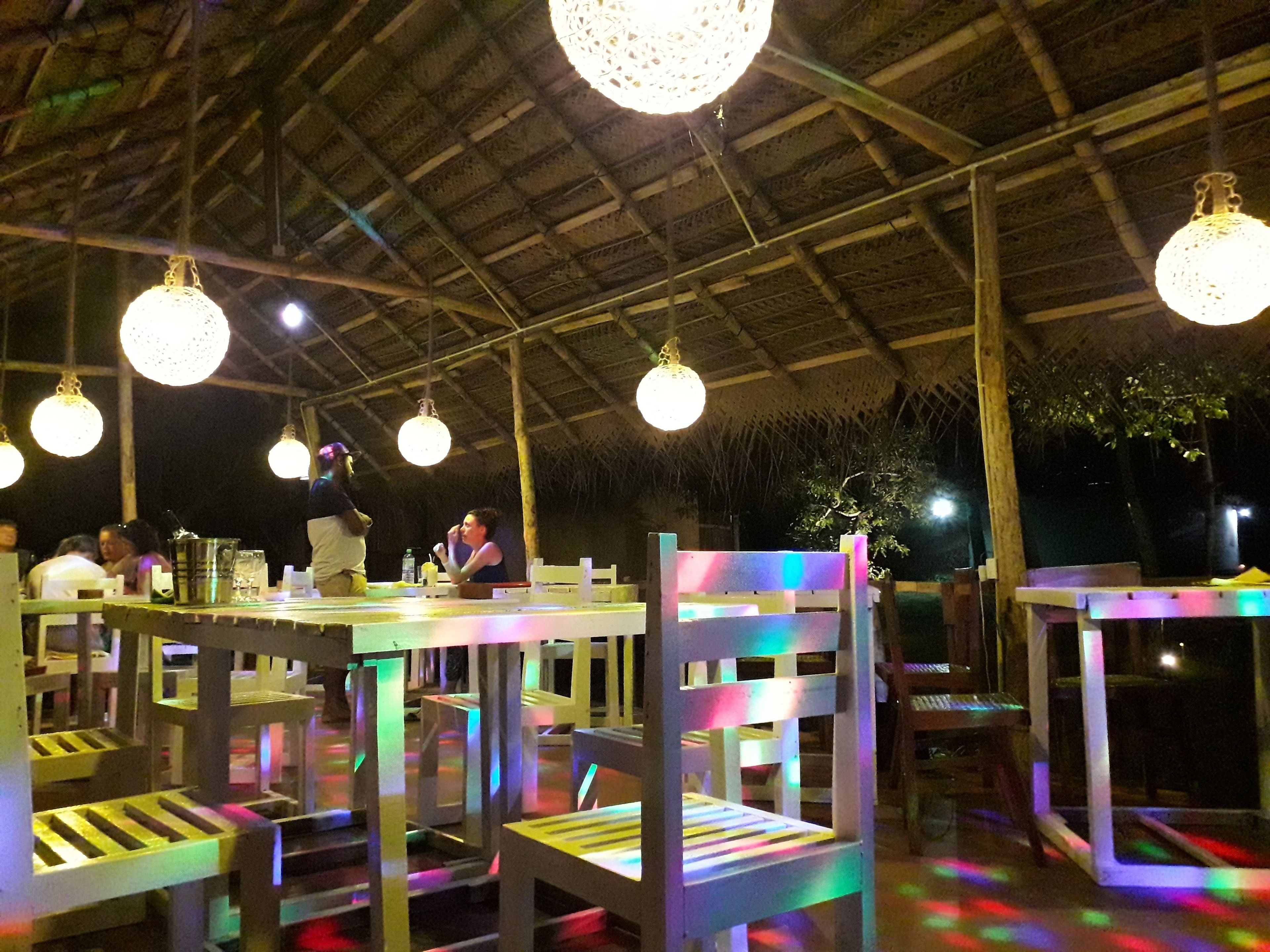 Een buitenterras met tafels en stoelen onder een rieten dak, verlicht met hangende bolvormige lampen.