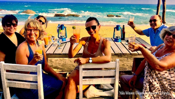 Een groep mensen zit aan een houten tafel op het strand, met de oceaan en rotsen op de achtergrond.